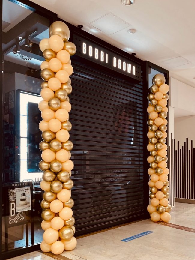 Balónková dekorace ve tvaru dvou balónkových sloupů z chromově zlatých a meruňkových balónků umístěných u chodu do Bobbi Brown prodejny
