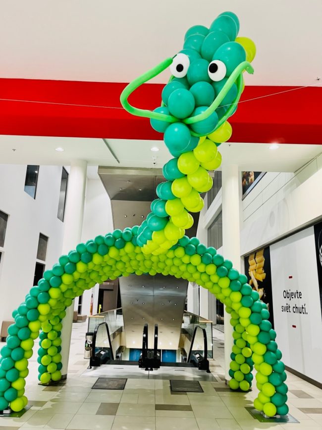 Dinosauří tunel z balónků u eskalátorů v obchodním centru, zelené balónky ve dvou odstínech