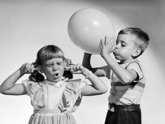 Pohled do historie balónků, černobílá fotka , chlapeček nafukuje balónek a holčička má zacpané uši.